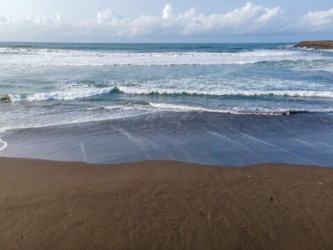 Aerial view of a dark sand beach with rolling waves, white foam, and a clear blue sky in Cilacap, Central Java, Indonesia.