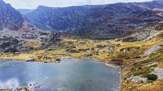 A lake sits in the foreground with rocky hills and grassy patches in the background. The sun shines down on the landscape, highlighting the rugged terrain during the day.