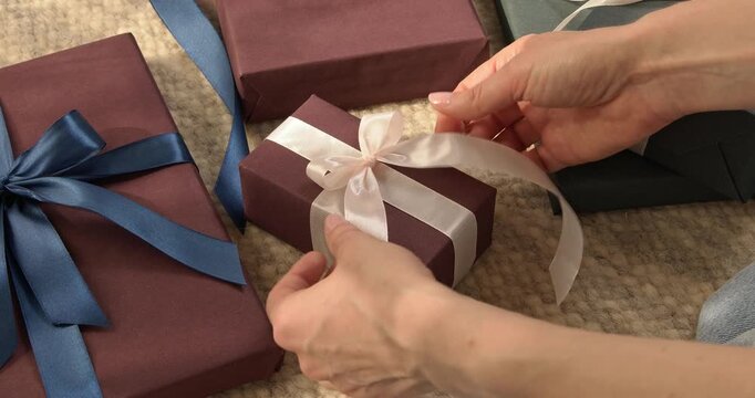 Woman hands forming bow from white satin ribbon on small gift box with other presents around close up video