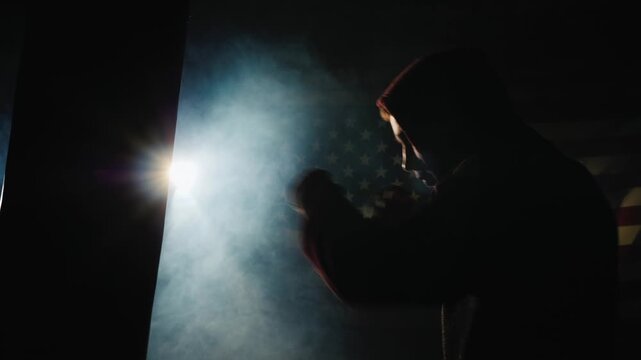 Silhouette of focused boxer training with punching bag in smoky dark gym with american flag.