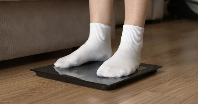 Woman feet in white socks stepping on digital electronic scales to check weight on wooden floor.