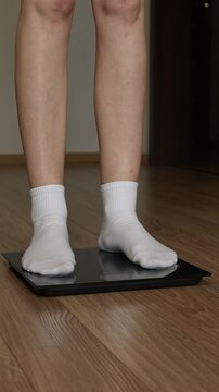 Woman feet in white socks stepping on digital electronic scales to check weight on wooden floor.