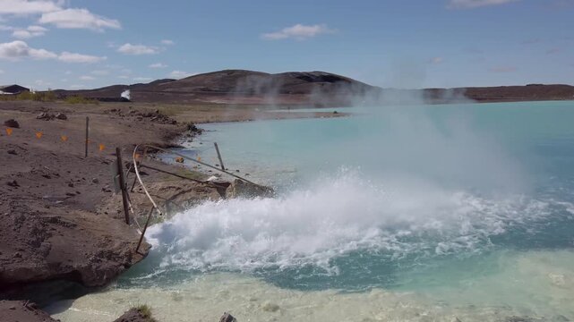 Geothermal outflow near Lake Myvatn, hot water gushing into a turquoise lake with spray and steam rising under a blue sky.