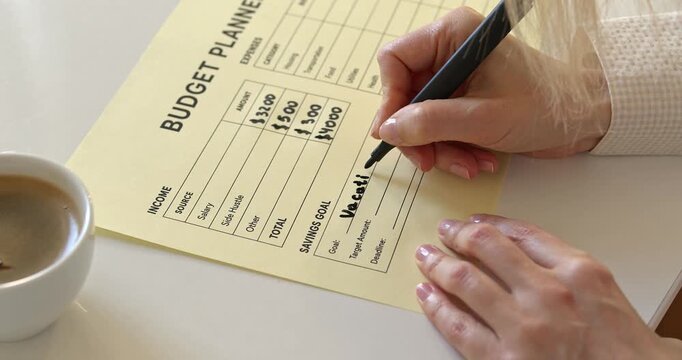 Woman hand writing vacation goal with black marker on paper budget planner next to coffee cup.