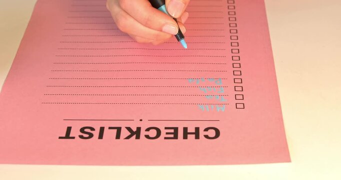 Close up of woman hand writing grocery checklist with blue marker on pink paper.