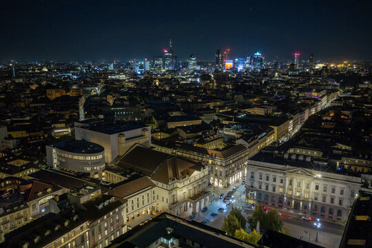 Milan, Italy - 23 September 2026: Aerial view of the illuminated La Scala opera house and the surrounding historic city center with the modern skyline glowing in the background at night.