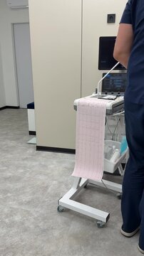 Medical cart with an ECG machine printing a pink electrocardiogram paper strip. Doctor in blue scrubs stands in a clinical hospital room with digital monitors.