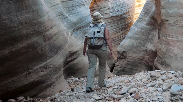 Woman with backpack walking through narrow smooth wavy slot canyon. Hiker touching layered sandstone walls and exploring scenic and remote wilderness of zion national park. Dried up riverbed