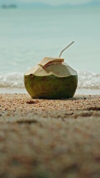 Fresh green coconut with drinking straw on tropical sandy beach with ocean waves in background.