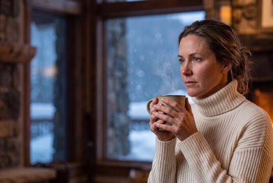 Thoughtful mature woman holding a steaming ceramic mug and looking out the window of a cozy rustic cabin in winter