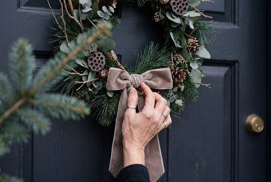 Close-up of a woman adjusting an elegant velvet bow on a handmade natural Christmas wreath hanging on a front door