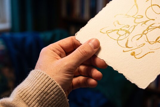 Close-up of a person holding handmade deckle-edge paper with ornate gold ink calligraphy lettering