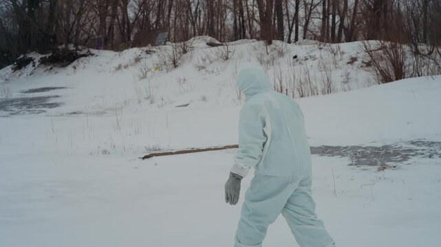 Researcher in white suit trudges along icy riverbank, scanning shore with gloved hand, bare trees and cracked ice in background, measured pace, focused survey in desolate winter landscape