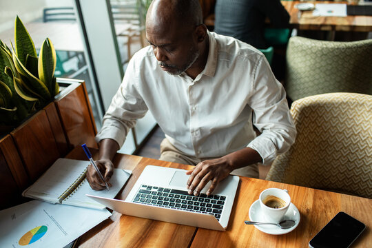 Man taking notes while working on laptop in cafe