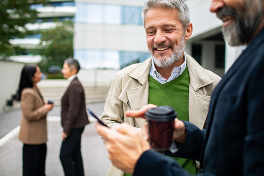 Senior businessman smiling at coworker?s smartphone while holding coffee
