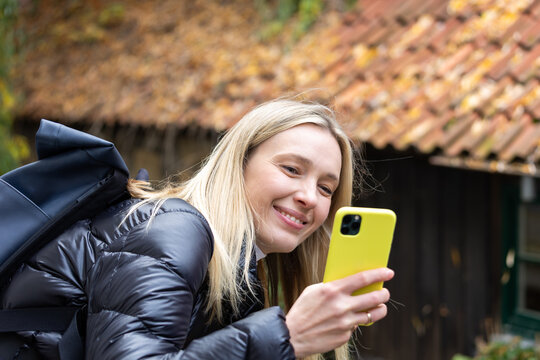 Young tourist woman smiles while using smartphone during autumn walk, carrying backpack near rustic buildings. Candid travel portrait shows cheerful traveler enjoying sightseeing and leisure outdoors