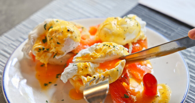 Delicious eggs benedict with smoked salmon and hollandaise sauce on a plate. Person cutting into a runny poached egg with a knife and fork. Gourmet breakfast meal in a restaurant setting.