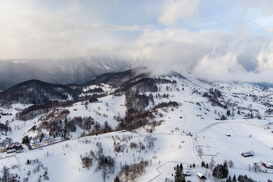 Brasov, Romania - 21 September 2026: Aerial view of the snow-covered Carpathian Mountains near Brasov, featuring rolling hills, scattered wooden cabins, and dense forests under a cloudy sky.