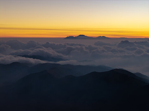 Aerial view of Mount Bromo at sunset, with golden light illuminating the horizon above a thick sea of clouds and dark mountain silhouettes in Tosari, East Java, Indonesia.