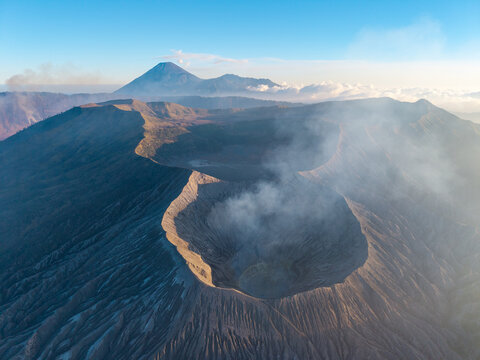 Aerial view of Mount Bromo crater with volcanic smoke rising from the rugged landscape under a clear blue sky in Tosari, East Java, Indonesia.