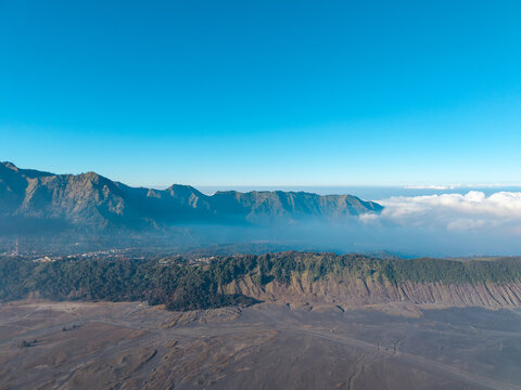 Aerial view of Mount Bromo landscape featuring rugged volcanic ridges, vast sandy plains, and low-hanging clouds under a clear blue sky in Tosari, East Java, Indonesia.