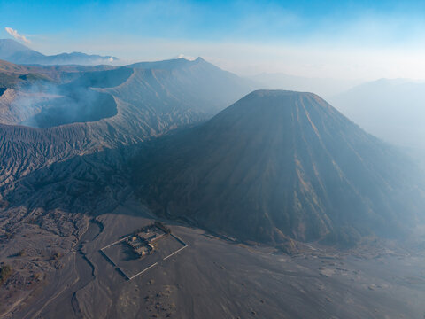Aerial view of the volcanic landscape of Mount Bromo with its iconic cone, smoking crater, and a small temple in the vast sandy caldera in Tosari, East Java, Indonesia.