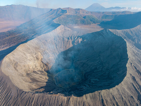 Aerial view of Mount Bromo crater with volcanic smoke, rugged ash-covered slopes, and surrounding mountain range under clear sky in Tosari, East Java, Indonesia.