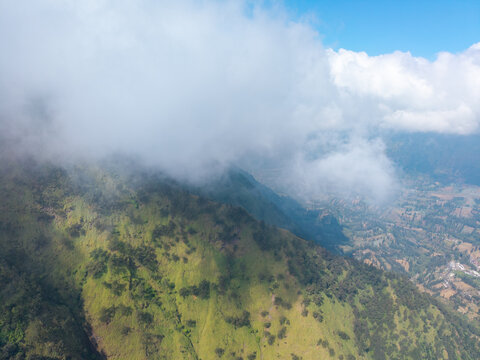 Aerial view of Wonokerto mountainside with lush green vegetation, scattered trees, and rolling hills partially obscured by soft white clouds in Sukorejo, East Java, Indonesia.
