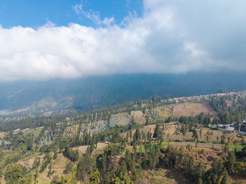 Aerial view of Wonokerto featuring terraced agricultural fields, lush green trees, and rolling hills under a cloudy sky in Sukorejo, East Java, Indonesia.