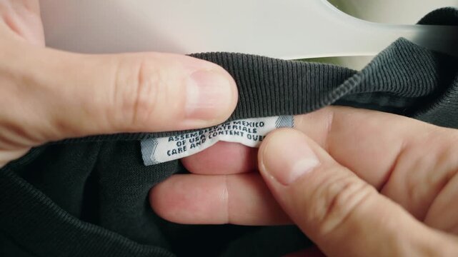 Close up of hands holding shirt label showing assembled in mexico of usa materials care and content over.