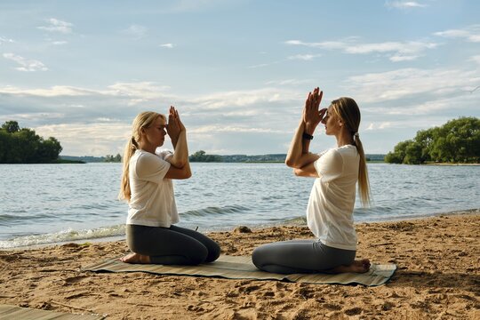 Two individuals are engaged in a yoga session on a sandy beach by a tranquil lake. The sun sets, casting soft colors across the sky. They perform a meditative pose sitting in front of each other