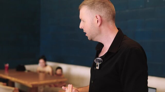 Young adult Caucasian man is standing in a empty cafe and explaining something gesturing his hands. Male influencer with lapel microphone on his shirt is communicating during the work brainstorm