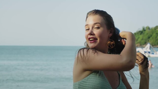 Pretty lady is standing on the beach and brushing her wet hair smiling and talking filmed at sea background. Cheerful young adult Caucasian woman in bikini is taking care of her coiffure on holidays