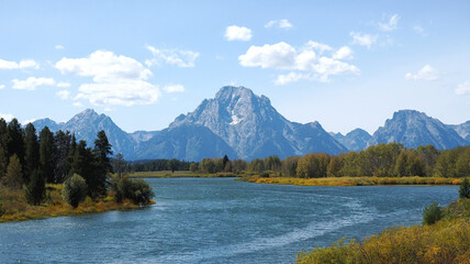 Serene river flows past towering mountain peaks. Grand Tetons, Oxbow Bend. Golden reeds line the water's edge. Evergreen trees frame the scene below. Clouds drift softly across the sky © Mark