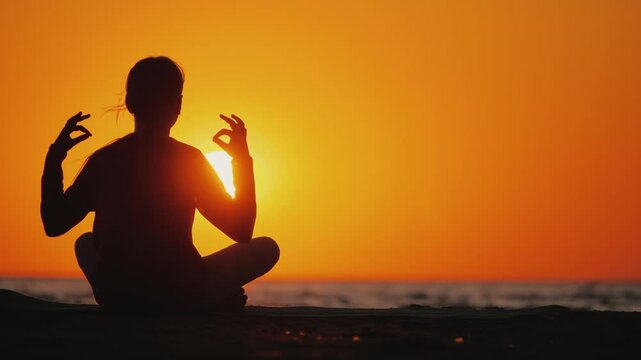 Silhouette of woman practicing yoga meditation in lotus pose on beach during golden sunset sky.