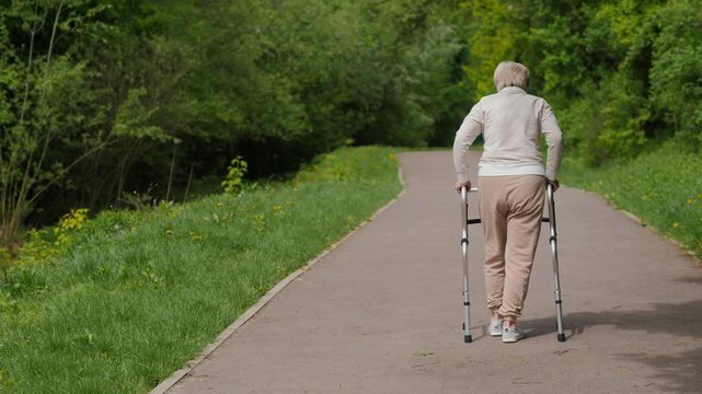 Elderly woman with walking frame on a paved path in a park, slow motion footage from behind.
