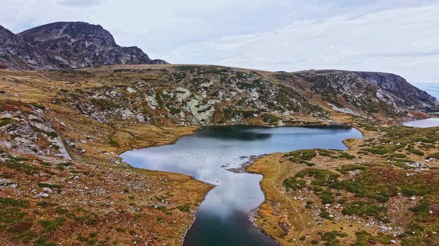 A large lake sits in the valley surrounded by mountains and rocky slopes. The landscape shows grass and stones with patches of greenery under a cloudy sky.