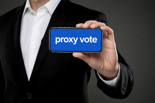 Businessman in black suit holds smartphone with proxy vote text on blue screen. Professional man shows mobile device for corporate voting shareholder decision. Digital concept of electronic election,
