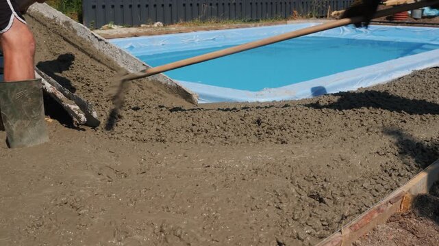 Two construction workers leveling wet concrete around outdoor swimming pool with rakes and shovels on sunny day.