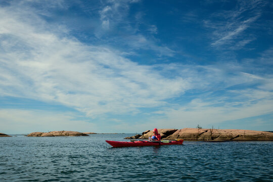 Solo sea kayaker resting in red kayak beside pink granite islands under wispy cirrus clouds, blue sky with copy space, Georgian Bay, Ontario