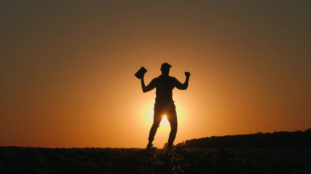 Silhouette of a man showing off his bicep muscles while holding a knife in a field at sunset.