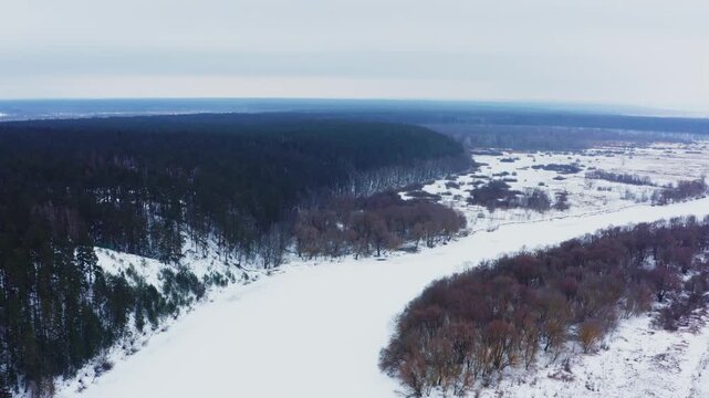 Drone flies over the snowy winter landscape with a frozen river. Beautiful nature in the winter. Aerial view of the snowy valley in winter.  Beautiful snowy landscape.