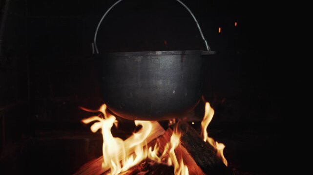 A close up slow motion video of black cast iron cauldron hanging over burning logs with fire sparks flying in the air at night in slow motion.