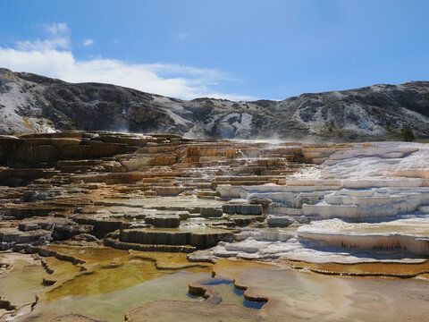 Yellowstone Mammoth Hot Springs stunning terraces glow with mineral hues. Steam rises from warm, sunlit pools below. Rugged mountains frame the vibrant, layered landscape. Crisp blue sky offers serene