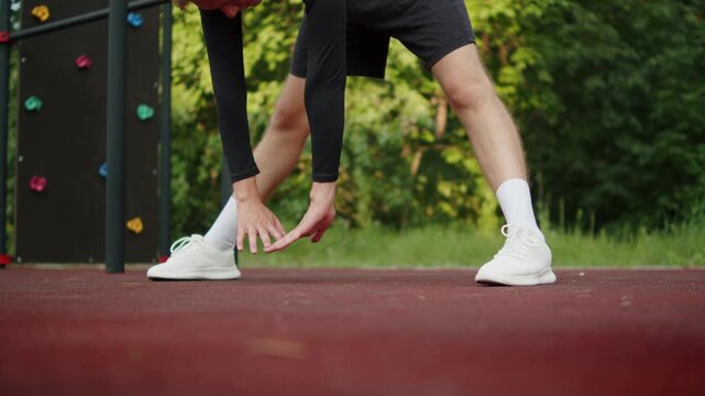 Athlete warming up on an outdoor training area. Man doing a wide-legged forward fold stretch. Sporty young man doing a forward bend stretch at an outdoor gym. Man on a sports groung.