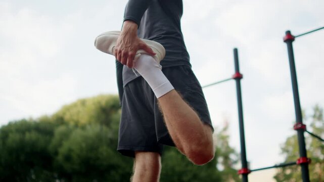 Man doing a standing leg stretch on an outside gym. Man in black athletic clothes pulling his foot to stretch his quadriceps muscle on an outdoor sports ground. Athlete training outdoors.