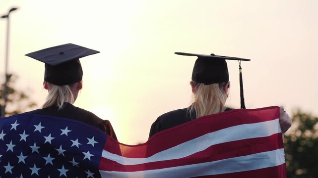 Two graduates in caps and gowns hold an American flag together raising their hands in triumph at sunset.