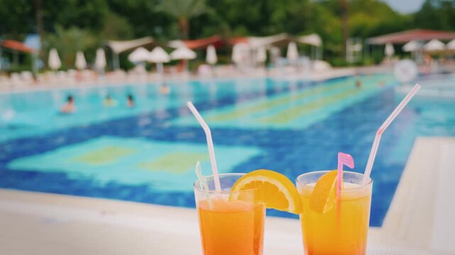 Two refreshing orange cocktails rest poolside with people swimming in background vacation footage.