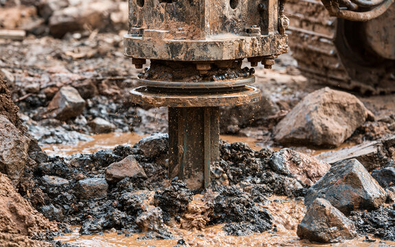 Close action shot of a large industrial drilling machine forcing its rotating drill into rocky underground layers, broken stones and dark wet soil displaced around the borehole, brown muddy water