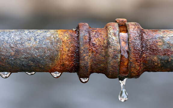 Corroded old pipe with rust texture shows dripping water drops. Metal joint connection point leaks liquid, indicating plumbing damage and system failure.